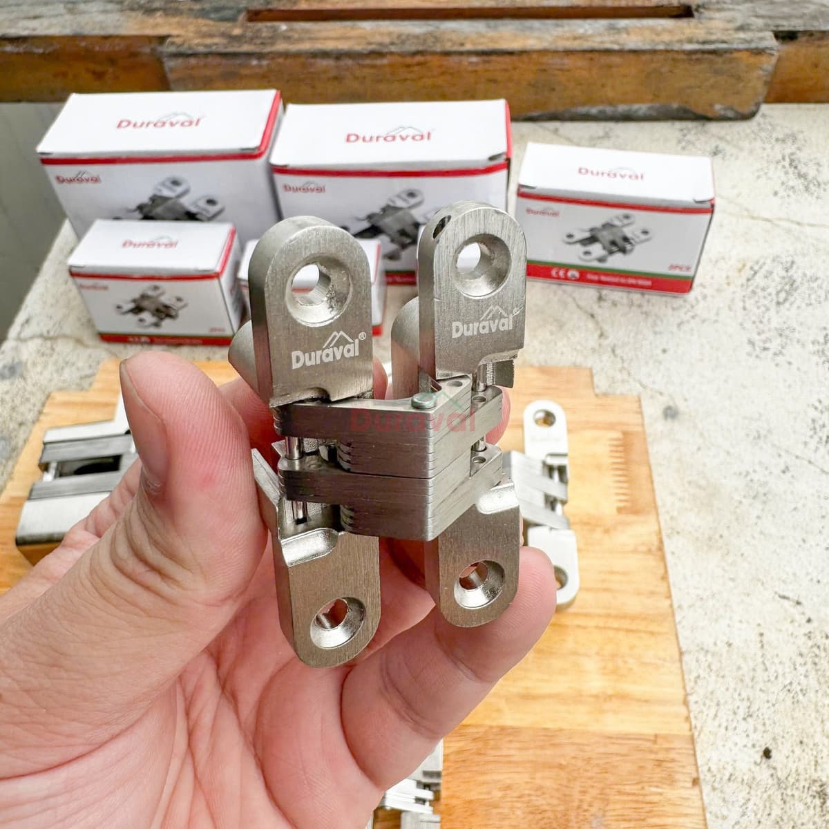Tay cầm bản lề âm chữ thập Duraval D201 có logo Duraval trên cánh bản lề, phía sau là các hộp sản phẩm