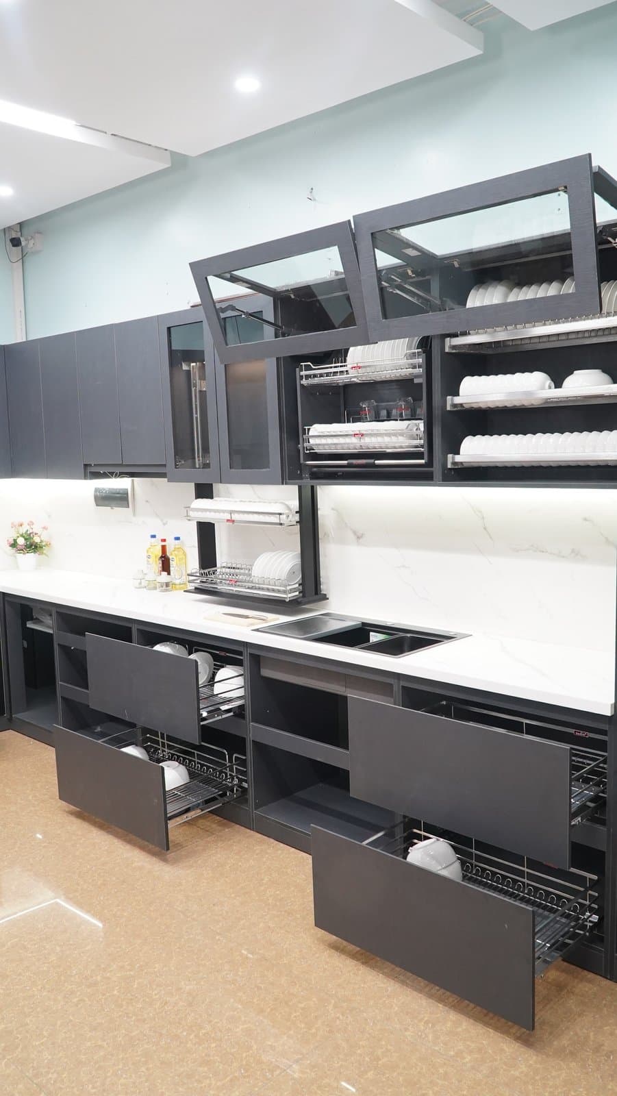 Bếp trưng bày Duraval tủ màu xám đen: tủ trên có giá bát inox, các ngăn kéo tủ dưới mở ra có giỏ inox đựng bát đĩa